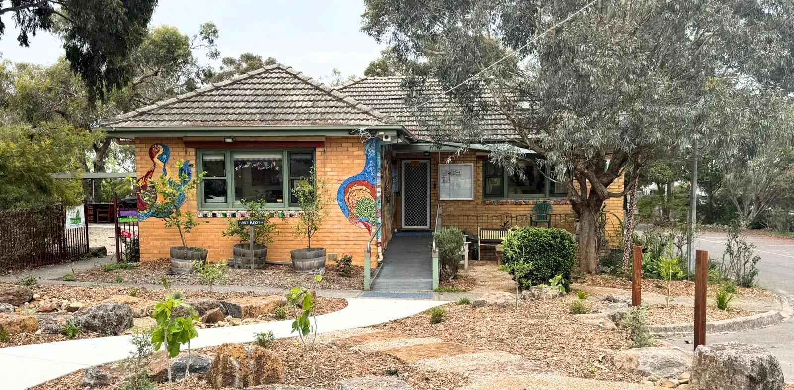 The neighbourhood house from the street, with a mosaic mural beside the door and a native garden out the front