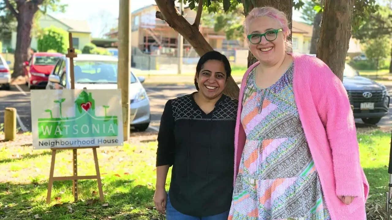 Two staff members standing together beside the Watsonia Neighbourhood House sign in the front garden