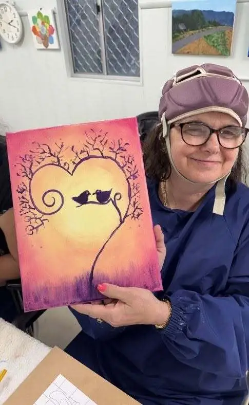 Rachel holding up a finished painting of two birds on a branch against a sunset