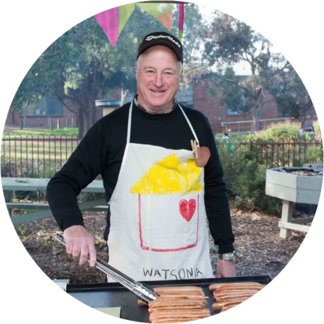 Bob Regan smiling in a Watsonia Neighbourhood House apron, cooking at the barbecue