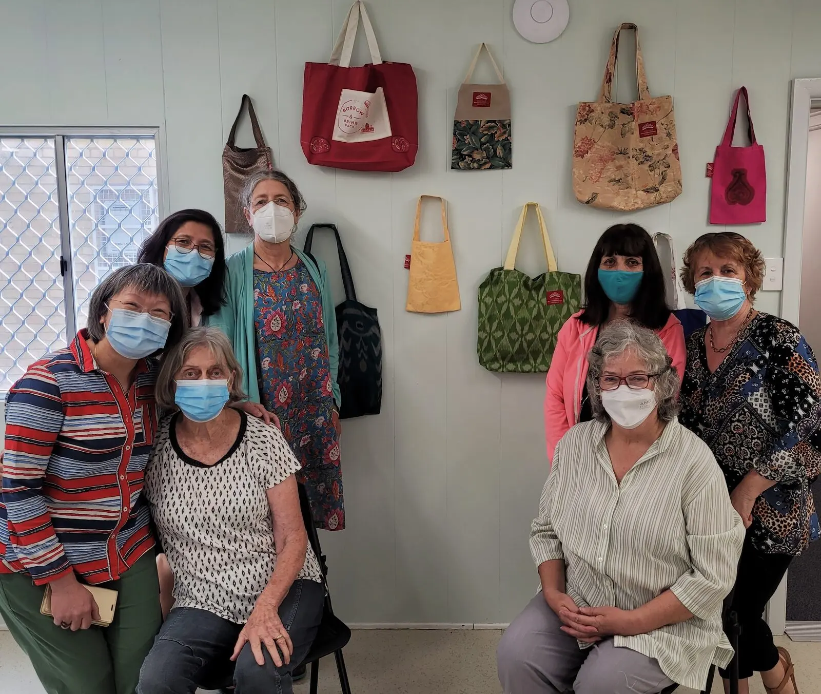 Six women from the Boomerang Bags group in front of a wall hung with the tote bags they have made
