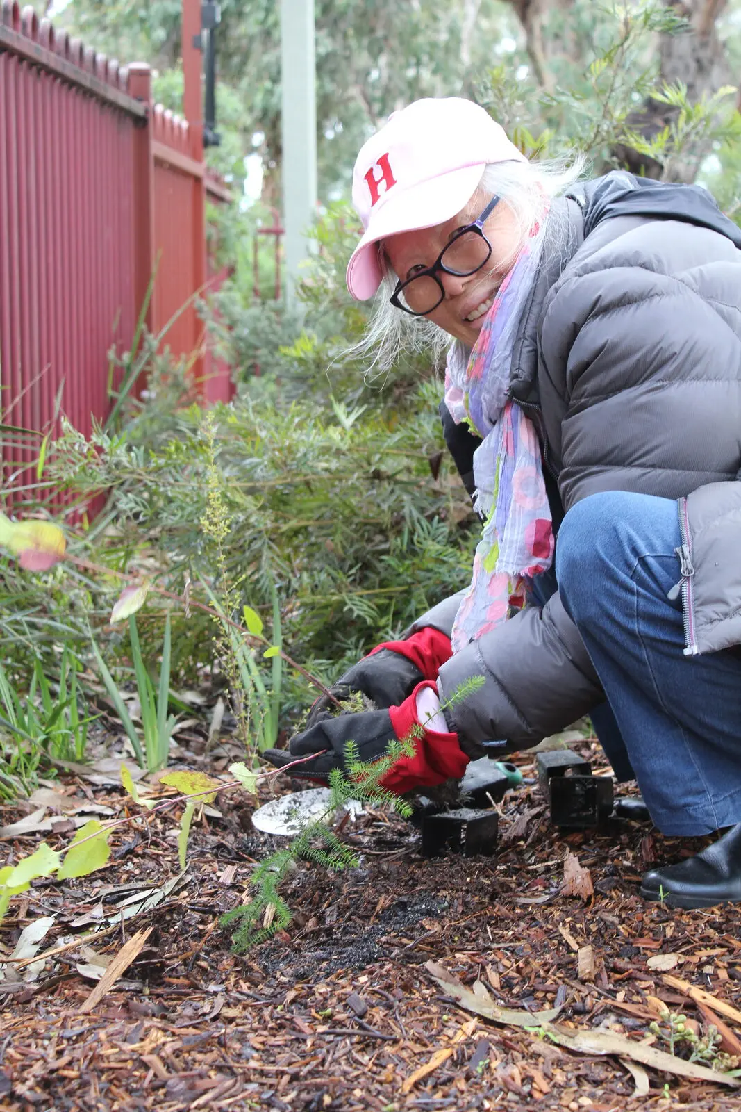 A volunteer in a cap and gloves planting into a garden bed beside the building