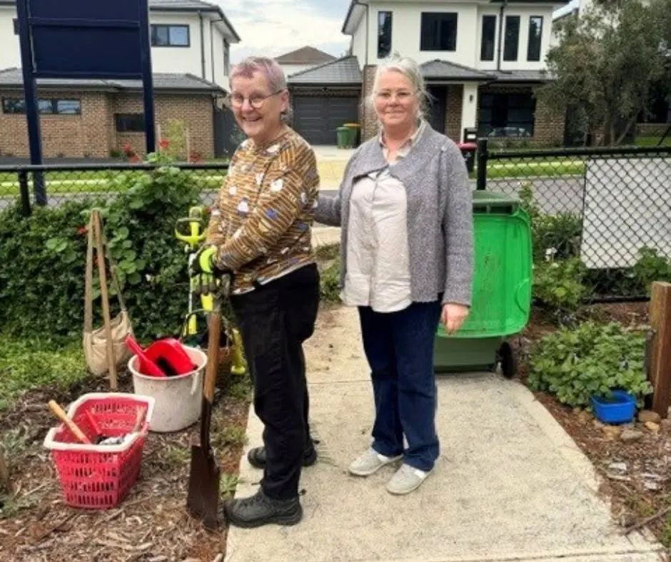 Two volunteers standing on the garden path beside a green bin, with buckets and garden tools at their feet