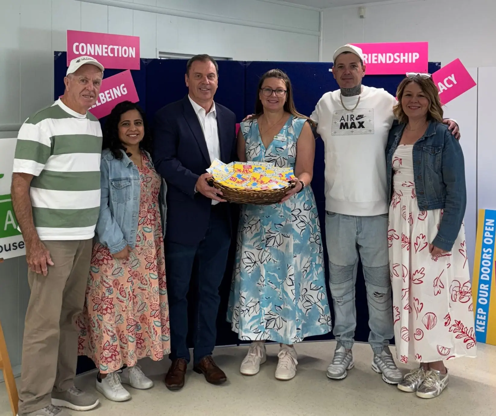 A group standing together indoors holding a platter, in front of a display of pink signs reading connection, wellbeing and friendship