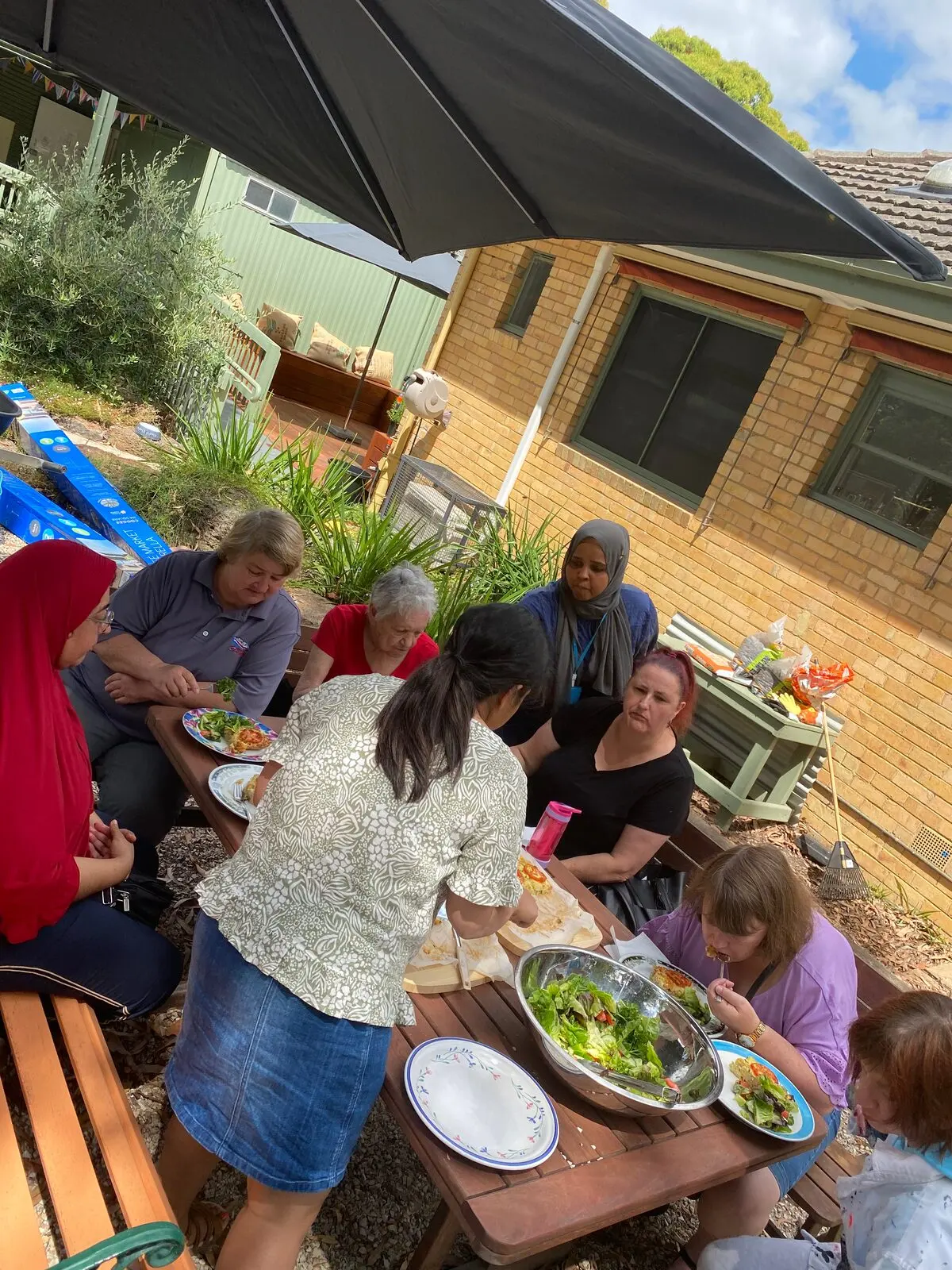 People serving themselves from platters at a long outdoor table beside the house