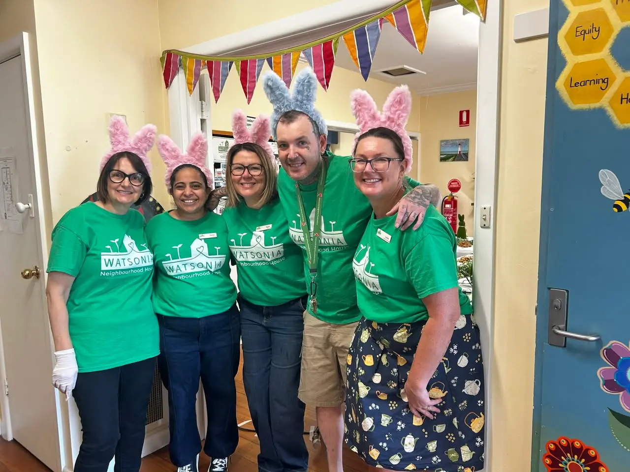 Five staff members in green Watsonia Neighbourhood House shirts and rabbit ears, standing together under bunting in the hallway