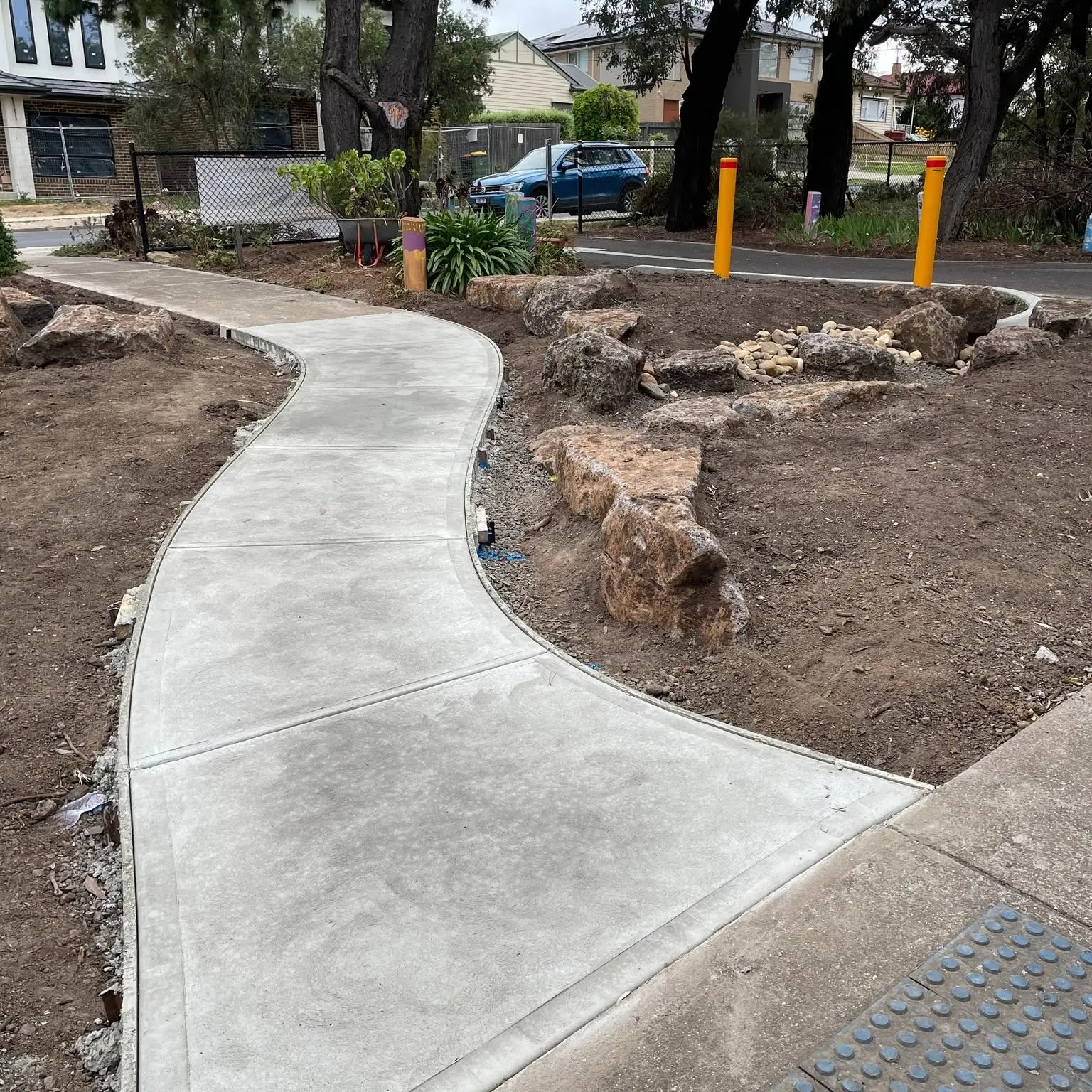 A curving concrete path winding through a new garden bed of rocks and young plants, with the street beyond
