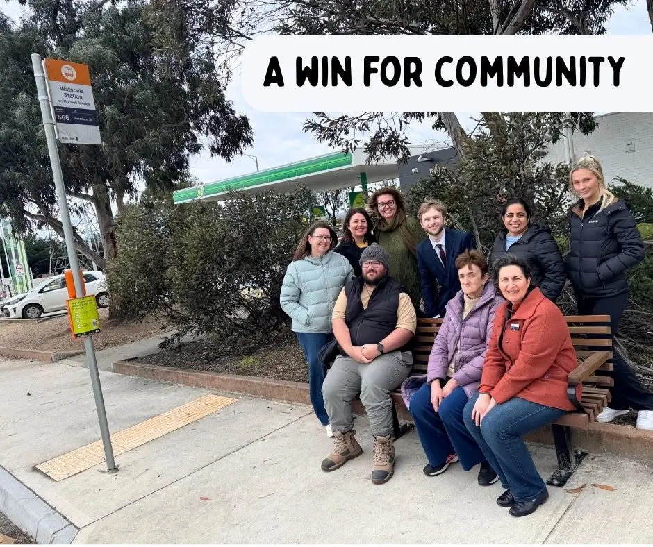 A group of people gathered around a new bench at a bus stop on Watsonia Road, with the bus stop sign and a service station behind