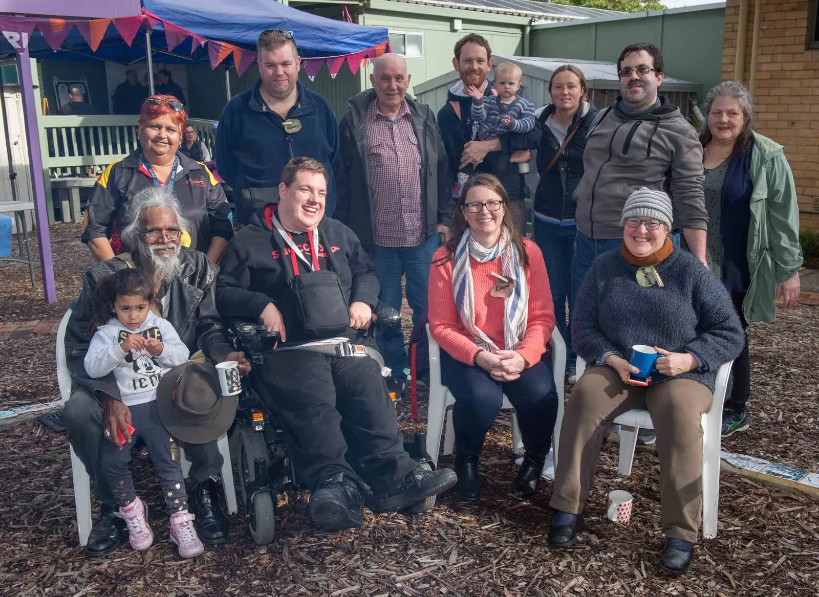 A dozen people of all ages gathered outdoors under bunting, several using wheelchairs