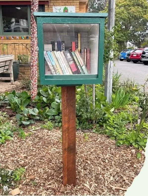 A green-framed little library box on a timber post in the front garden, its glass door showing a shelf of books
