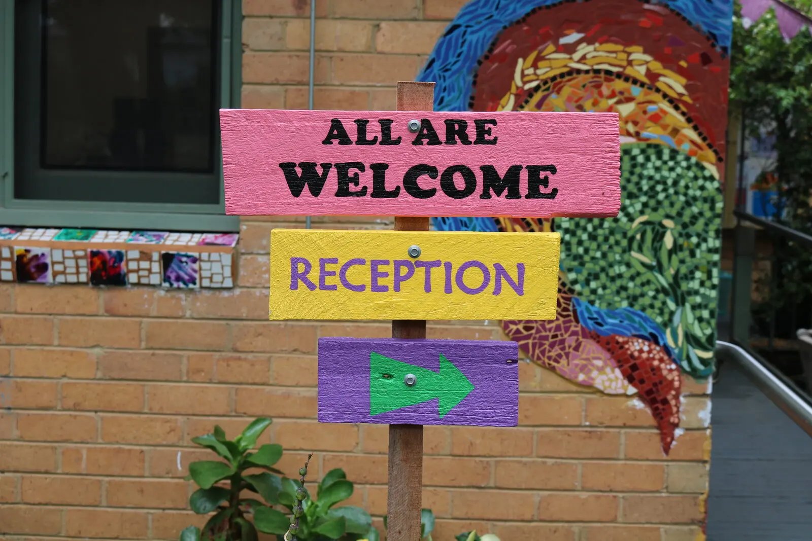 Hand-painted signs on the brick wall beside the door reading All Are Welcome and Reception, next to a bright mosaic mural